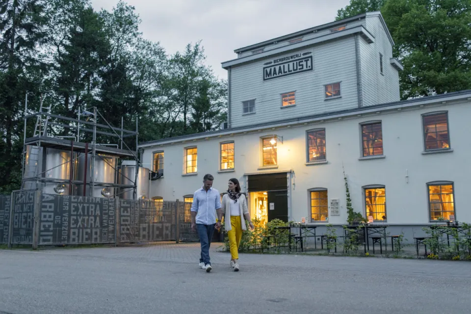 Verlicht wit gebouw van brouwerij Maallust in Veenhuizen met bezoekers op het plein ervoor