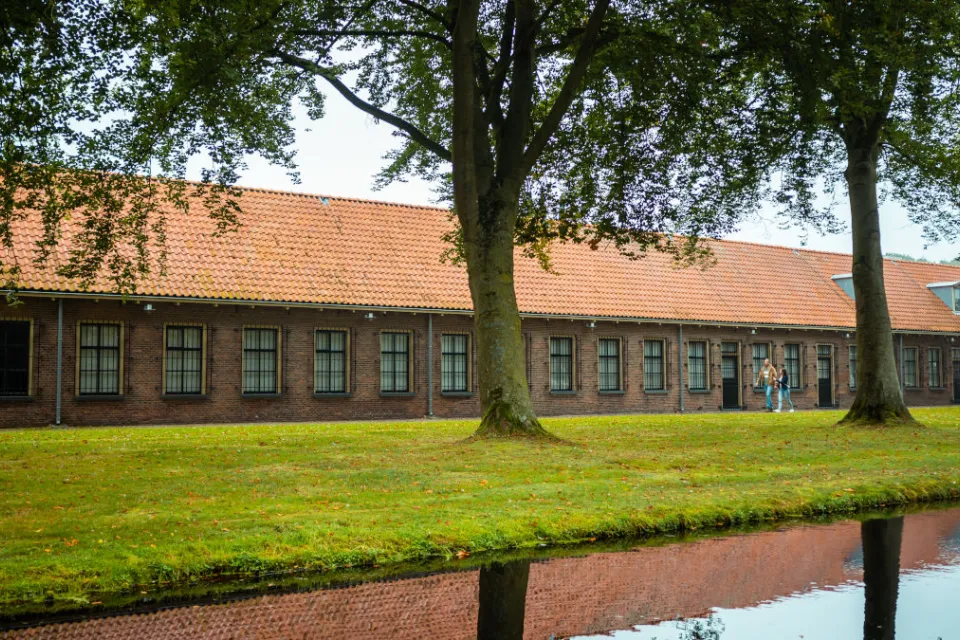 Langgerekt bakstenen gebouw met rode daken aan een vaart met bomen in Veenhuizen