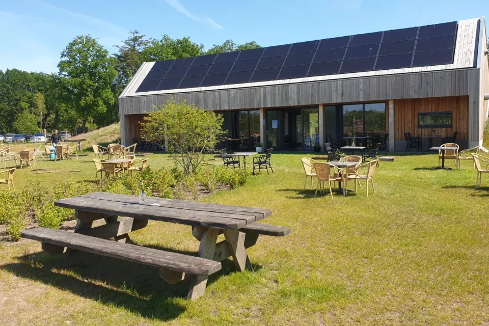 Modern houten bezoekerscentrum met zonnepanelen, terrasstoelen en een picknicktafel aan de bosrand
