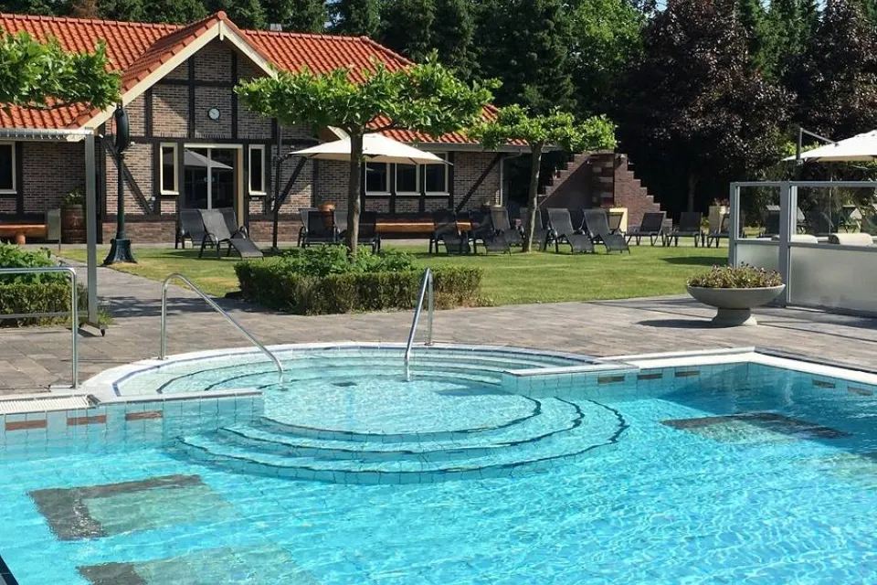 Verwarmd buitenbad van een spa met ligstoelen, parasols en een houten gebouw op de achtergrond