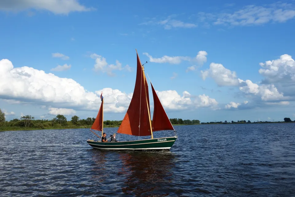 Zeilboot met rode zeilen vaart over een groot meer onder een blauwe lucht met wolken